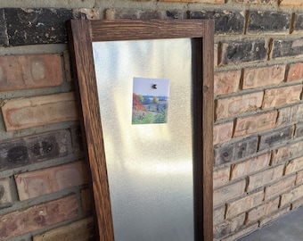 May include: A rectangular magnetic board with a dark brown wooden frame. The board has a silver metal surface with a small photo attached by a magnet. The photo shows a landscape scene with trees and a body of water.