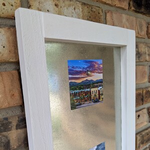 May include: A white framed magnetic board with two photos attached. The top photo shows a cityscape at sunset with mountains in the background. The bottom photo shows a rocky landscape with a cloudy sky.