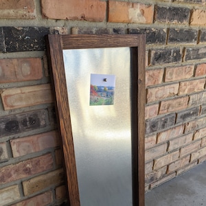May include: A rectangular magnetic board with a dark brown wooden frame. The board has a silver metal surface with a small photo attached by a magnet. The photo shows a landscape scene with trees and a body of water.