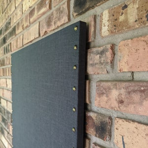 May include: A dark gray fabric bulletin board with gold tacks around the edge, mounted on a brick wall.