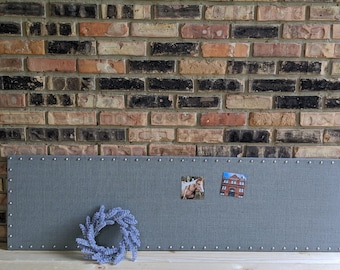May include: A gray fabric memo board with silver nailhead trim, two small pictures, and a lavender wreath. The board is rectangular and mounted against a brick wall. The pictures are of a horse and a building.