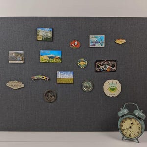 May include: A large rectangular dark gray fabric-covered magnetic bulletin board featuring various colorful souvenir magnets depicting locations like Detroit, Iowa, and Las Vegas, accompanied by a vintage-style light blue twin-bell alarm clock.
