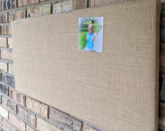 May include: A beige burlap bulletin board with a photo of a child pinned to it. The board is mounted on a brick wall.