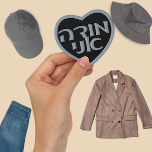 May include: A black heart-shaped patch with the text "Drink JIC" in white lettering. The patch is held in a hand against a beige background.