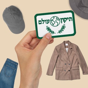 May include: A green and white embroidered patch with the text "מִיקוֹד עֲלַם" and a globe graphic. The patch is being held by a hand.