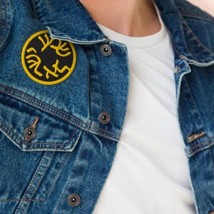 May include: A blue denim jacket with a circular black patch displaying yellow Hebrew letters. The patch is attached to the left breast. The jacket has a traditional design with button fastenings and two chest pockets.