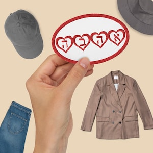 May include: A red oval patch with four white hearts containing Hebrew letters. The patch is being held by a hand against a beige background.