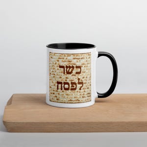 May include: A white ceramic mug with a black handle and interior rim. The exterior features a square graphic of matzah bread with the Hebrew text "כשר לפסח" printed in brown letters across the centre of the textured, light brown cracker pattern.