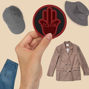May include: A red embroidered patch with a black background and a hand with the Hebrew word "Hamsa" in the centre. The patch is being held by a hand.