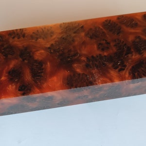 May include: A rectangular block of wood with a unique, swirling pattern of dark brown and orange hues. The wood has a smooth, polished finish.