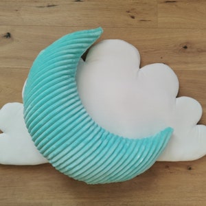 Op de afbeelding: Een turquoiseblauw halvemaanvormig kussen op een wit wolkenvormig kussen. Beide kussens zijn gemaakt van zacht, pluche stof.