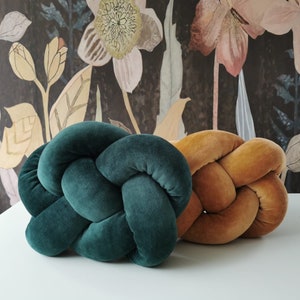 Może przedstawiać: Dwie splecione poduszki, jedna w ciemnozielonym aksamicie, a druga w ciepłym brązowym aksamicie. Poduszki są ułożone na białej powierzchni.