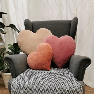 May include: Three heart-shaped pillows in shades of pink, peach, and beige are arranged on a gray armchair with a black and white chevron patterned throw blanket.
