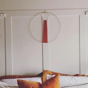 May include: A white wall with a gold metal hoop and a red wooden dowel hanging on it. The wall is panelled and has a white bed with brown and white pillows in front of it.