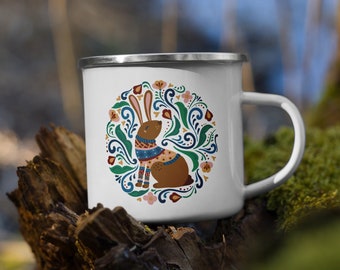 Könnte beinhalten: Weiße Email Tasse mit einem braunen Kaninchen, das einen blau-weißen Pullover trägt, in einem Kreis aus grünen Blättern, rosa Blumen und blauen Schnörkeln.