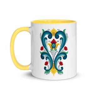K&ouml;nnte beinhalten: Eine wei&szlig;e Keramiktasse mit gelbem Rand und einem blauen und gelben Blumendesign. Das Design zeigt eine stilisierte Blume mit roten Akzenten.
