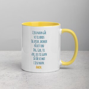 K&ouml;nnte beinhalten: Eine wei&szlig;e Keramiktasse mit gelbem Rand und Henkel. Die Tasse zeigt ein blaues Textdesign mit der Aufschrift "I JESU NAVN G&Aring;R VI TIL BORDS OG SPISER, DRIKKER P&Aring; DITT ORD DEG, GUD, TIL &AElig;RE, OSS TIL GAVN S&Aring; F&Aring;R VI MAT I JESU NAVN. AMEN."