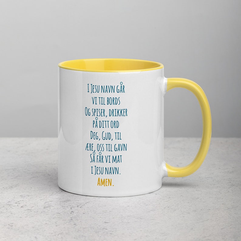 K&ouml;nnte beinhalten: Eine wei&szlig;e Keramiktasse mit gelbem Rand und Henkel. Die Tasse zeigt ein blaues Textdesign mit der Aufschrift "I JESU NAVN G&Aring;R VI TIL BORDS OG SPISER, DRIKKER P&Aring; DITT ORD DEG, GUD, TIL &AElig;RE, OSS TIL GAVN S&Aring; F&Aring;R VI MAT I JESU NAVN. AMEN."
