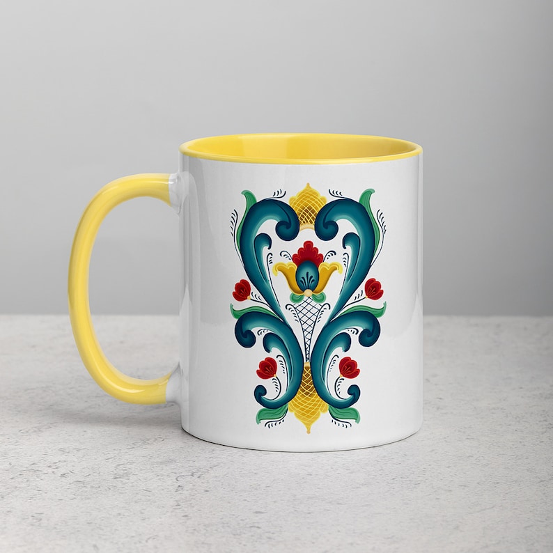 K&ouml;nnte beinhalten: Eine wei&szlig;e Keramiktasse mit gelbem Rand und einem blauen und gelben Blumendesign. Das Design zeigt eine stilisierte Blume mit roten und gelben Akzenten.