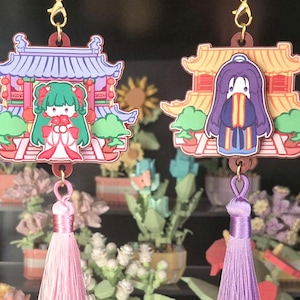 Peut inclure: Deux pendentifs décoratifs avec des personnages de dessins animés dans des bâtiments de style asiatique. Chaque pendentif a un gland, un rose et un violet. Les pendentifs sont suspendus à des crochets dorés. Le fond présente des plantes en pot miniatures.