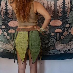 May include: A handmade green crochet skirt with a brown waistband and pointed edges. The skirt is worn with a gold-colored tank top. The background features a tapestry with a forest and mushroom design.