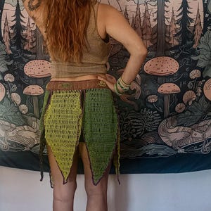 Puede incluir: Una falda de ganchillo verde hecha a mano con una cinturilla marrón y bordes puntiagudos. La falda se lleva con una camiseta sin mangas de color dorado. El fondo presenta un tapiz con un diseño de bosque y setas.