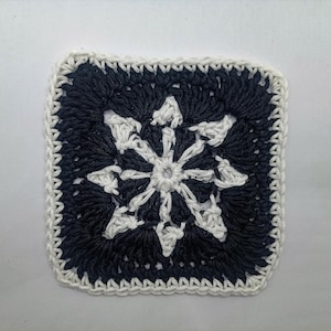 May include: A square, crocheted piece featuring a central white snowflake design against a navy blue background. The edges are finished with a white border, creating a contrasting frame. This handmade item showcases intricate stitch work.