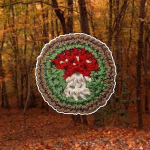 May include: A round, crocheted coaster featuring a mushroom design. The mushroom has a red cap, a white stem, and is surrounded by green and brown yarn. The coaster is set against a blurred background of a forest with autumn colors.