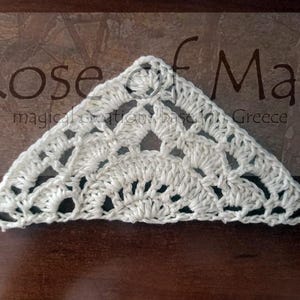 May include: White crocheted triangle with a lacy design. The crochet piece is on a brown wooden surface. The text "Rose of May" is visible in the background.