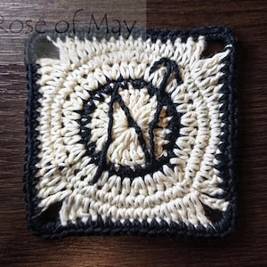 May include: A crocheted square with a black border and a white center. The center has a design that looks like a letter N.