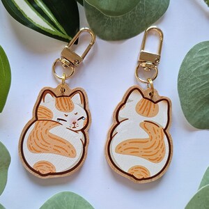 Könnte beinhalten: Zwei Katzen-Schlüsselanhänger aus Holz. Jeder Schlüsselanhänger zeigt ein Cartoon-Katzen-Design mit weißen und orangefarbenen Markierungen. Die Schlüsselanhänger haben goldfarbene Verschlüsse zum Befestigen an Schlüsseln oder Taschen. Das Gesicht der Katze ist auf einer Seite sichtbar, und der Rücken der Katze ist auf der anderen Seite sichtbar.