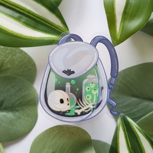 Könnte beinhalten: Ein transparenter Rucksack aus Plastik mit einem Schädel, Knochen und einem Glas mit Augen darin. Der Rucksack ist mit grüner Flüssigkeit gefüllt und hat eine Fledermaus auf der Vorderseite.