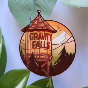 Könnte beinhalten: Ein runder Aufkleber mit dem Schriftzug "GRAVITY FALLS" in Braun. Das Design zeigt einen Wasserturm, Berge und Kiefern vor einem Sonnenuntergangshimmel. Der Aufkleber ist von grünen Blättern umgeben.