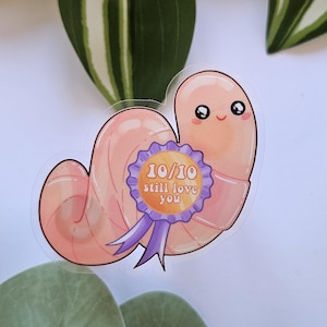 Puede incluir: Una pegatina caprichosa con un gusano de dibujos animados con una cara sonriente y una insignia que dice "10/10 still love you". El gusano es de color melocotón con una insignia morada y dorada. Fondo blanco con hojas verdes.