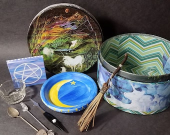May include: A collection of witchy items including a round box with a green and blue chevron pattern, a round box with a painted scene of a forest and a moon, a small blue and white tile with a pentacle, a small blue and yellow plate with a crescent moon and a star, a glass bowl, two silver spoons, a small black knife, and a broom.