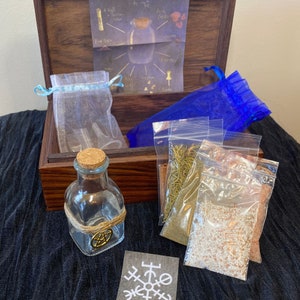 Puede incluir: Una caja de madera con una bolsa de cord&oacute;n azul y blanco, una botella de vidrio transparente con un tap&oacute;n de corcho y un amuleto de pentagrama dorado, y cuatro peque&ntilde;as bolsas de pl&aacute;stico transparente que contienen hierbas y especias secas. La caja tiene una receta escrita a mano en la tapa interior.