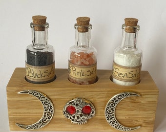 Puede incluir: Tres pequeños frascos de vidrio con tapones de corcho llenos de sal negra, sal rosa del Himalaya y sal marina. Los frascos están dispuestos en un soporte de madera con un diseño de calavera y media luna.