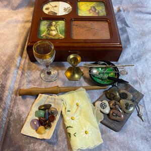 May include: A wooden box with four compartments, each containing a different image. The box is surrounded by various items, including a glass goblet, a small gold candlestick, a bowl with a green and black design, a wooden wand, a small bag of crystals, a small bag of stones with symbols, and a yellow handkerchief with white daisies.