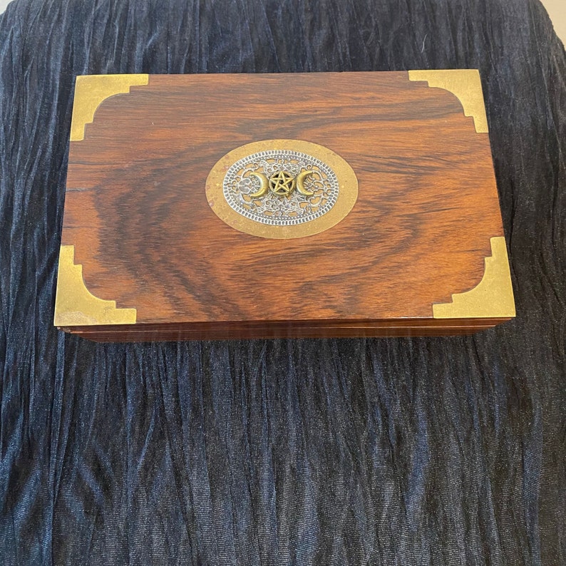 Puede incluir: Una caja de madera con ribete de lat&oacute;n y un c&iacute;rculo met&aacute;lico decorativo con un s&iacute;mbolo de triple luna en el centro.