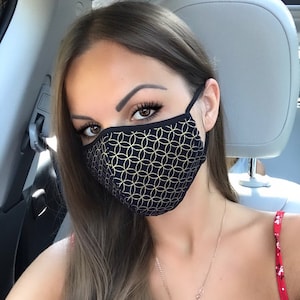 Pode incluir: Uma mulher usando uma máscara facial preta com um padrão geométrico dourado. A máscara cobre o nariz e a boca. Ela está usando um colar de ouro com um pingente.