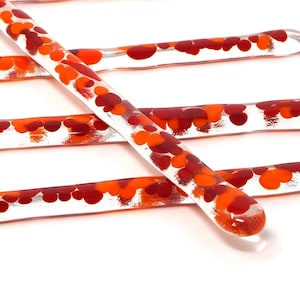 Könnte beinhalten: Fünf klare Glasrührstäbchen mit roten und orangen Flecken. Die Stäbchen sind kreuzweise angeordnet.