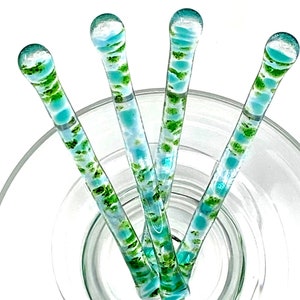 May include: Four glass stirring sticks with a blue and green swirl pattern. The sticks are arranged in a fan shape and are sitting in a clear glass bowl.