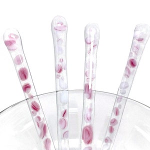 May include: Four glass stirring sticks with pink and white swirls. The sticks are clear glass with pink and white swirls throughout. The sticks are in a clear glass bowl.