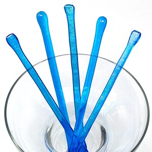 Op de afbeelding: Vijf blauwe glazen roerstokjes in een helder glazen martini-glas. De stokjes zijn waaiervormig gerangschikt.