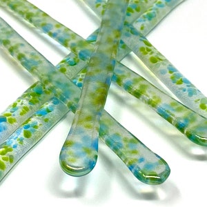 Puede incluir: Primer plano de varios palitos para remover de vidrio transparente con un patrón moteado azul y verde. Los palitos están dispuestos en un patrón entrecruzado sobre un fondo blanco. Los extremos son redondeados.