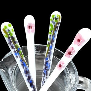 Könnte beinhalten: Vier weiße Glasrührlöffel mit Blumendesigns. Die Löffel befinden sich in einem durchsichtigen Glaskrug mit Henkel.