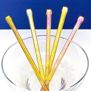 Op de afbeelding: Vijf iriserende glazen roerstokjes in een martini-glas. De stokjes zijn goud en roze met een glinsterende afwerking.