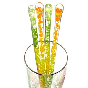 May include: Four clear glass stir sticks with colourful orange and green speckled designs. The stir sticks are displayed in a clear glass. The stir sticks have a rounded top and are straight. The glass has a gold rim.