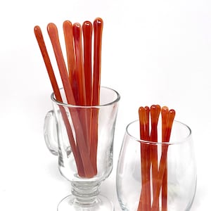 Pode incluir: Dois recipientes de vidro cheios de vários palitos de agitação de vidro laranja. Os palitos são de dois tamanhos diferentes e estão dispostos verticalmente nos recipientes.