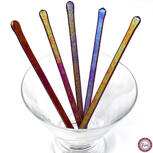 Op de afbeelding: Vijf iriserende glazen roerstaafjes in een heldere martiniglas. De staafjes hebben een platte, rechthoekige vorm met afgeronde uiteinden. De kleuren verschuiven van goud naar paars en blauw. Het glas is helder en de achtergrond is wit.